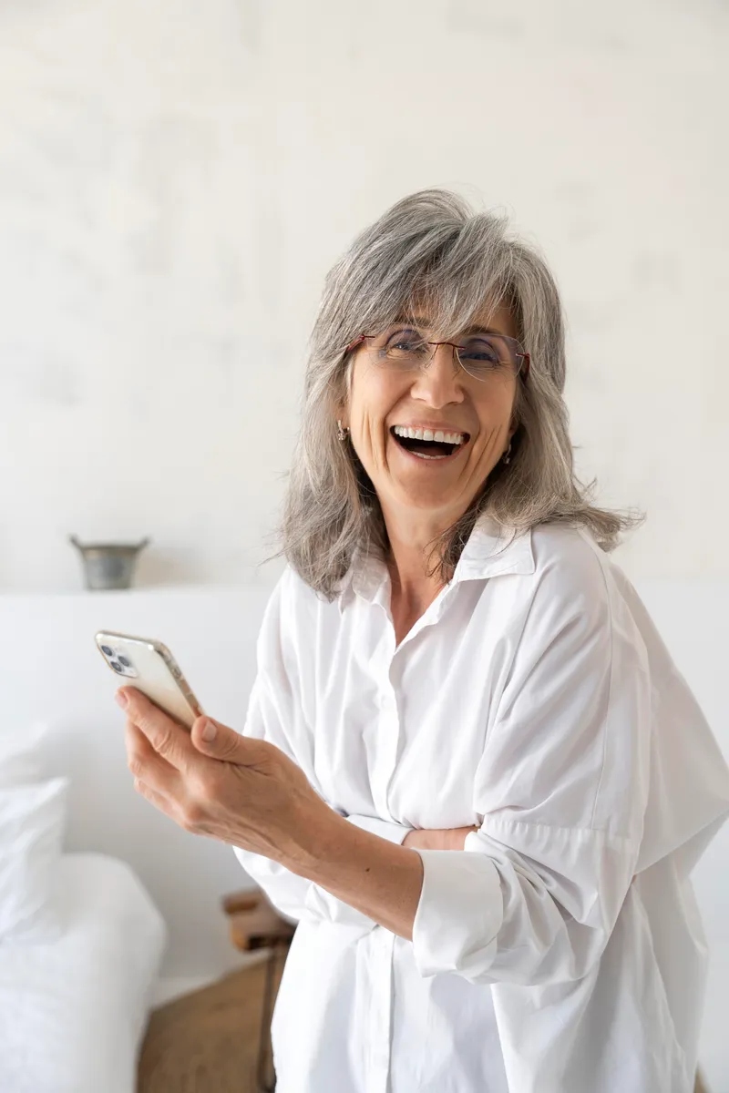 Aposentada rindo enquanto usa o celular em casa
