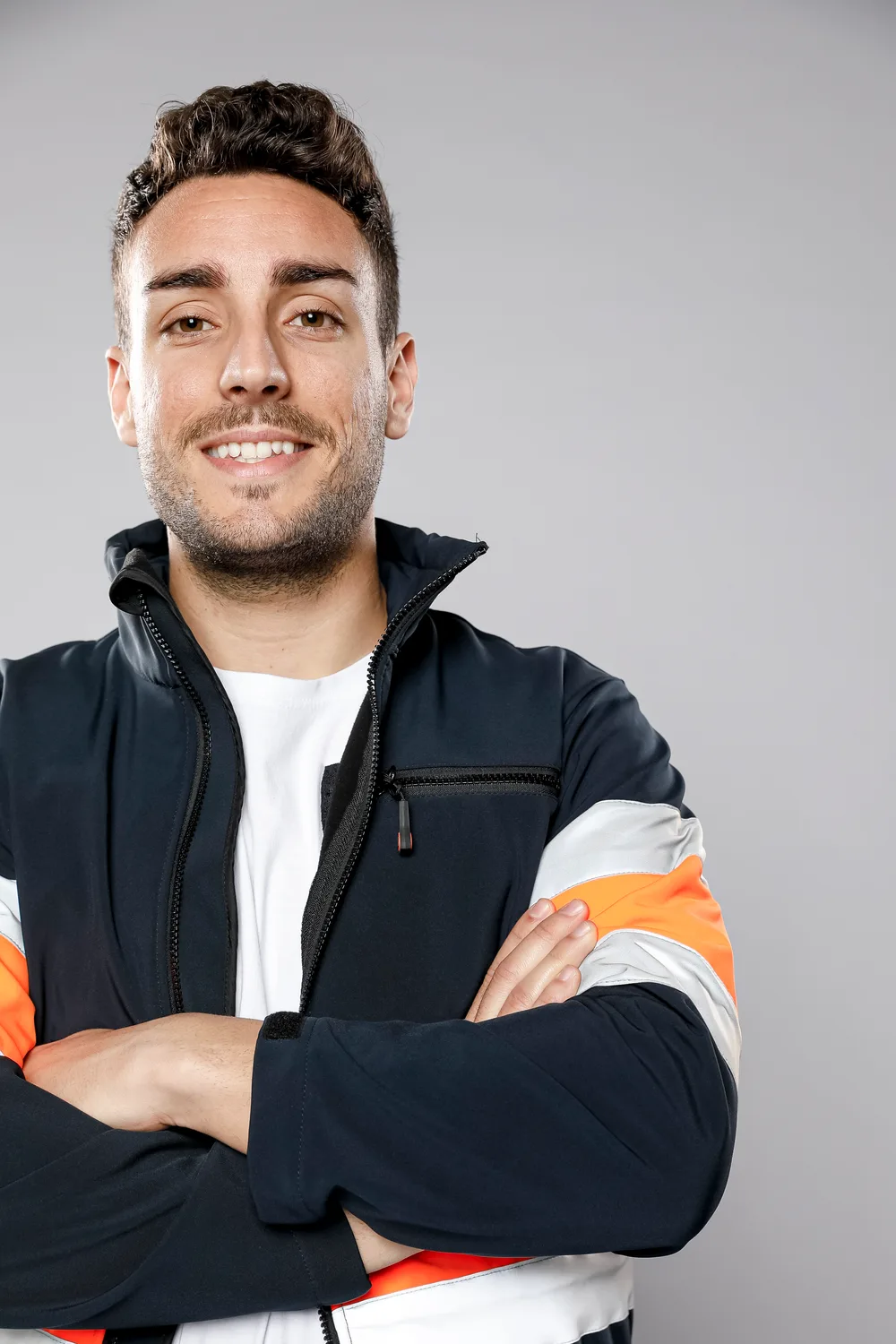 Jovem trabalhador sorrindo de braços cruzados, com jaqueta de trabalho